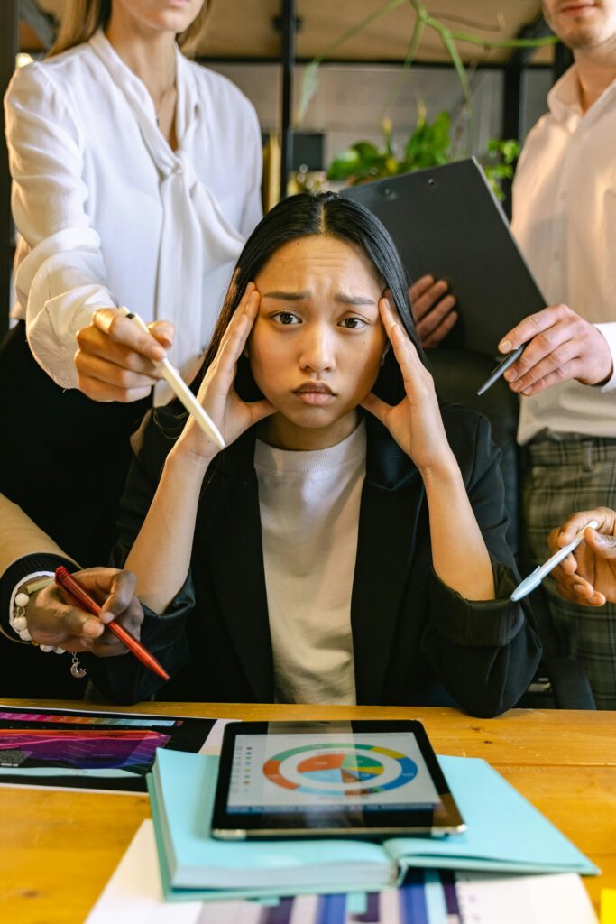 A stressed young woman overwhelmed with work at the office, surrounded by colleagues and devices.
