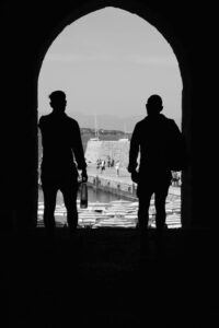 Two silhouetted figures framed by an archway gaze over Cefalù's historic harbor at sunset.