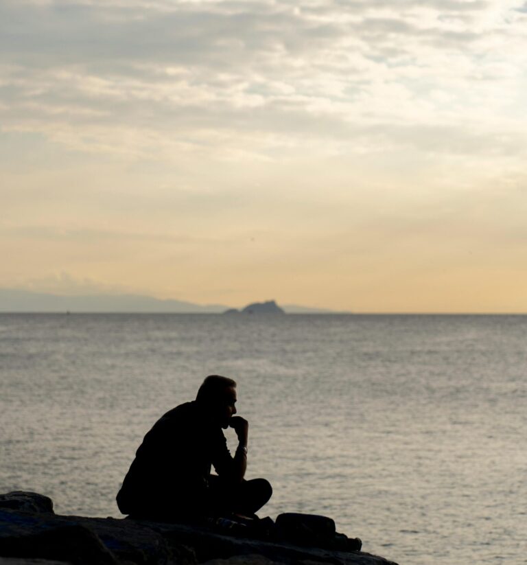 Silhouette of a person sitting by the sea during sunset, evoking a sense of contemplation and tranquility.